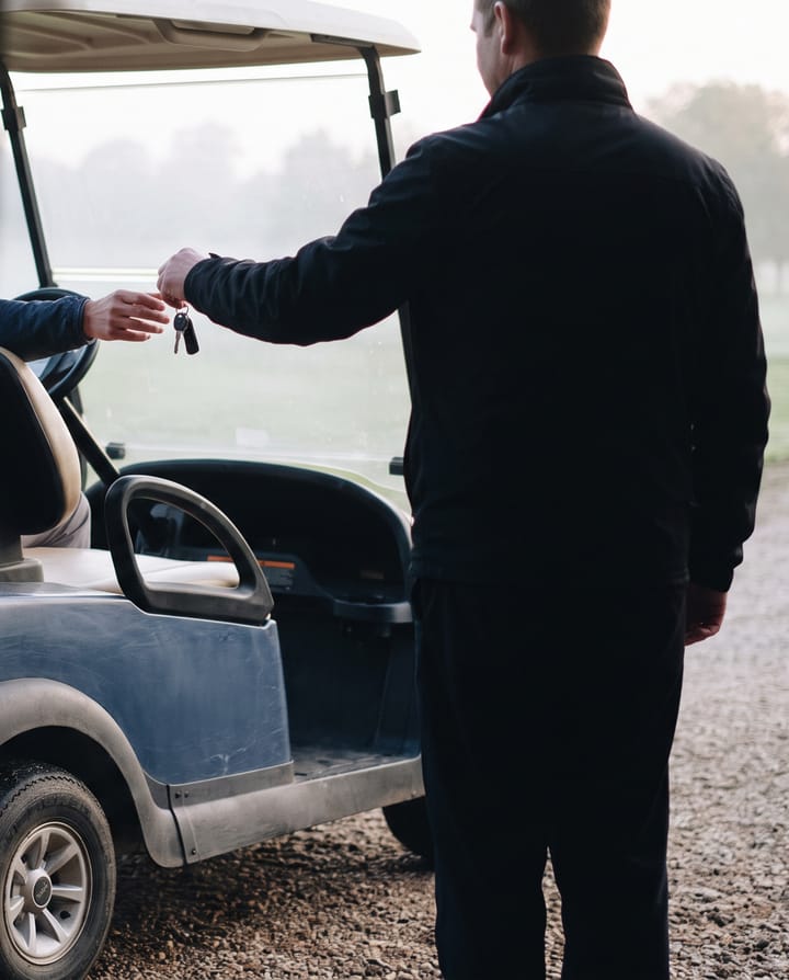 An attendant seen from behind handing keys across to a player beside a golf cart pulled up at a stone clubhouse door, the fairway and morning mist beyond.
