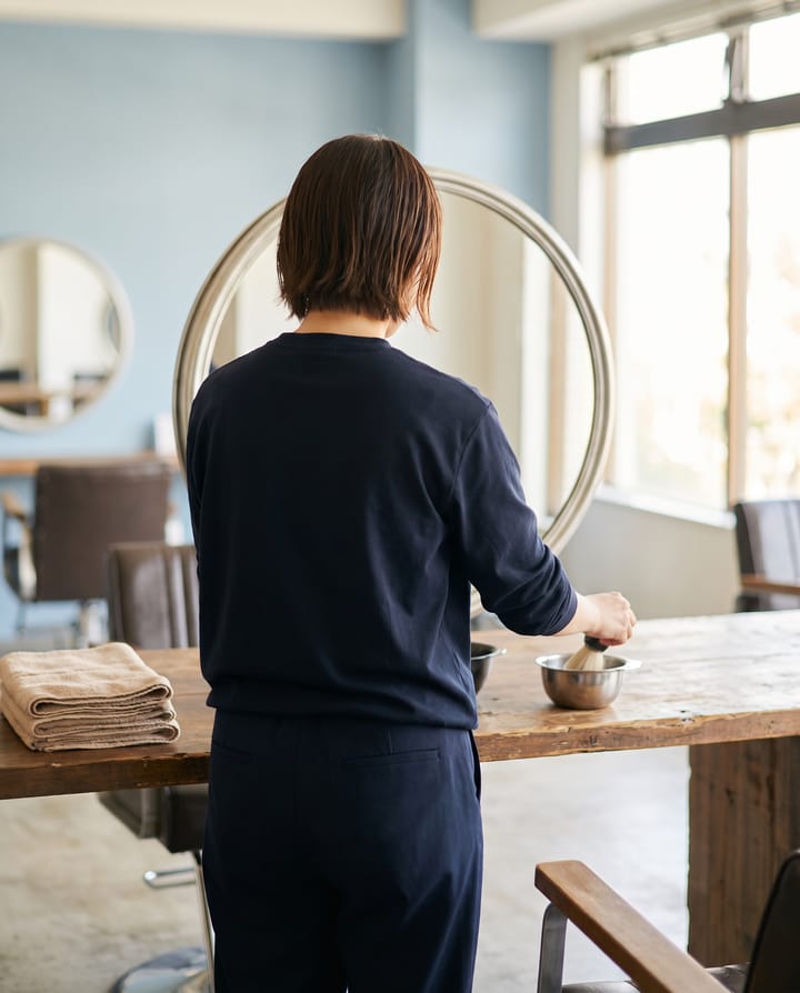 A salon station at opening: a chair facing a round mirror, a bowl and brush and folded towels on a warm wood counter, a stylist seen from behind setting out tools by a window.